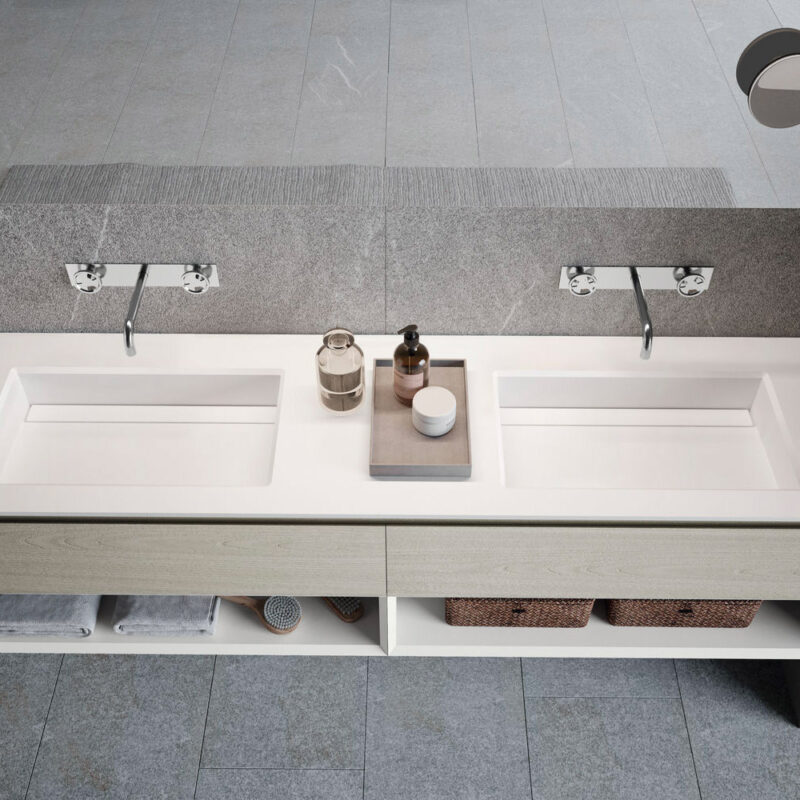 Vista dall'alto di un ampio mobile lavabo sospeso con doppio lavello integrato in resina bianca opaca. La struttura presenta frontali in legno chiaro con vano a giorno inferiore organizzato con asciugamani e cestini, completata da due rubinetti a parete cromati su parete effetto pietra grigia.