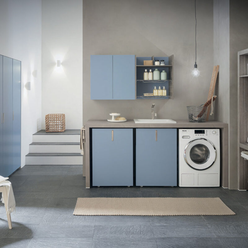 Panoramica di un'ampia e luminosa lavanderia moderna organizzata sui toni dell'azzurro polvere e del grigio. La composizione centrale include un piano da lavoro grigio con lavatoio incassato, una lavatrice a vista e basi con ante azzurre, sormontato da pensili abbinati. Sulla destra si trova una cabina armadio a vista in legno grigio con cesti e grucce, mentre a sinistra è visibile un armadio a muro azzurro.