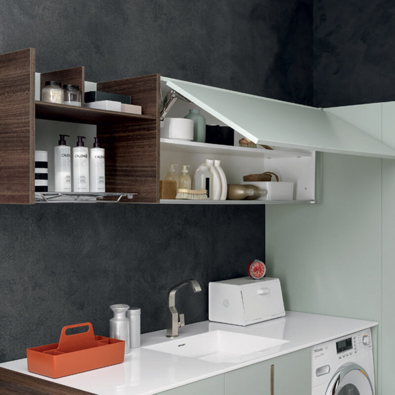 Vista di pensili da bagno e lavanderia a parete aperti. Sulla sinistra si trova un modulo a giorno in legno scuro con flaconi di sapone, mentre sulla destra un pensile orizzontale in finitura verde salvia/acquamarina chiarissimo con anta a vasistas sollevata mostra ripiani interni organizzati con accessori per la cura personale.