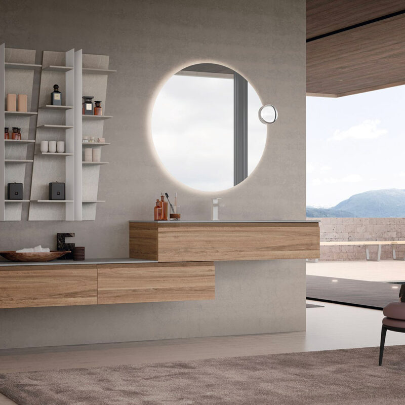 Composizione bagno moderna con mobili sospesi sfalsati in legno naturale, specchio circolare retroilluminato con specchietto ingranditore e modulo pensile a giorno con mensole, ambientata in una stanza luminosa con vista panoramica.