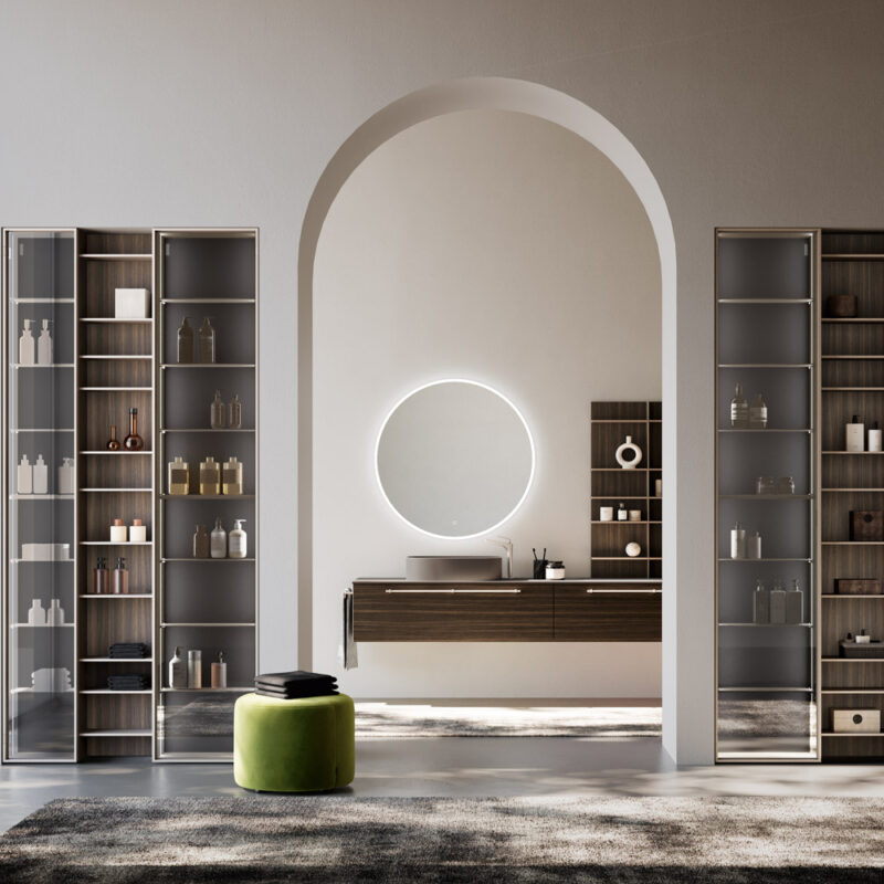 Bagno moderno di design con lavabo sospeso in legno scuro, specchio rotondo retroilluminato e scaffali con ante in vetro. Ambiente luminoso con arco centrale e sgabello verde su tappeto scuro.