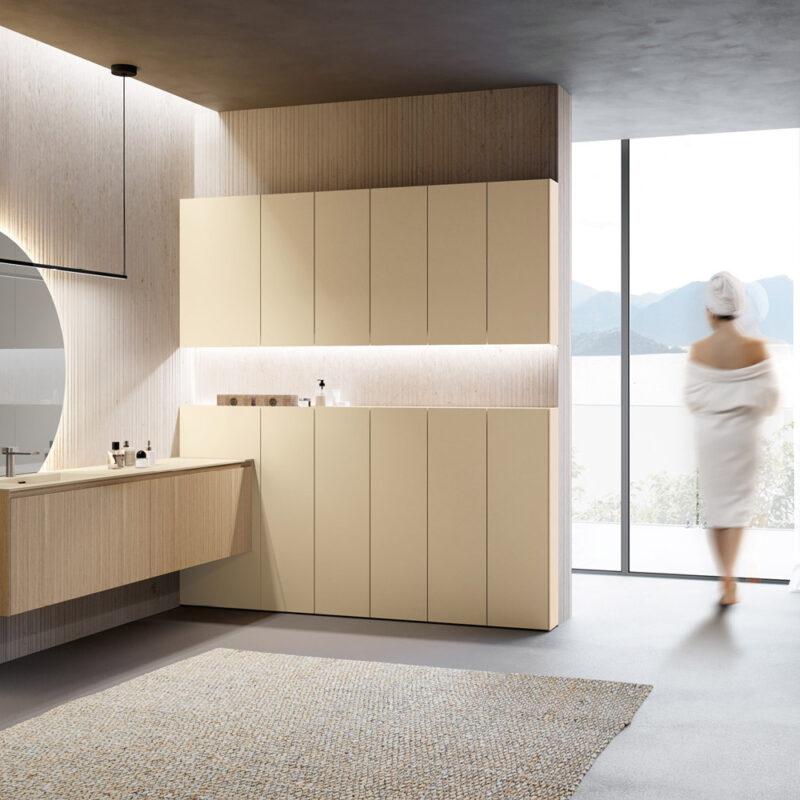 Bagno moderno con mobili sospesi della collezione Lime 2.0 di Azzurra e colonne contenitive color beige, grande specchio rotondo retroilluminato, luce naturale e donna in accappatoio di spalle.