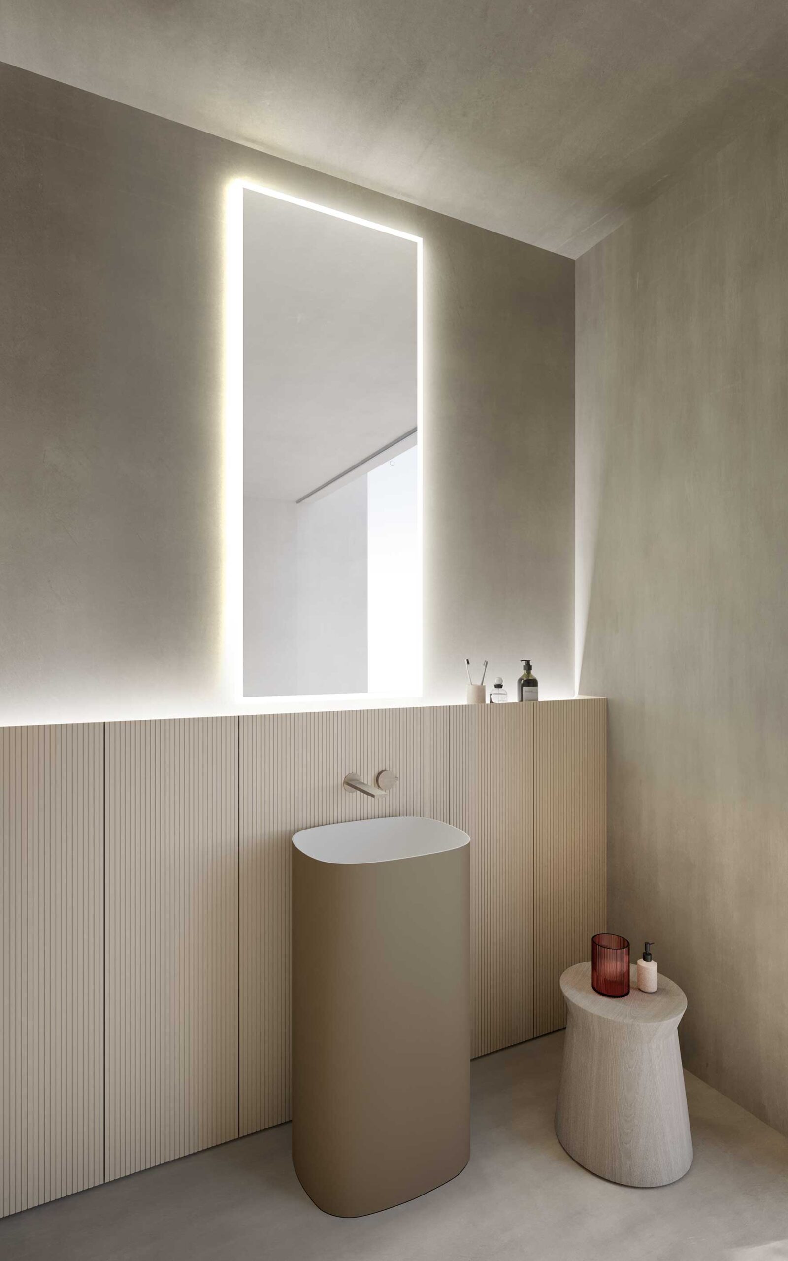 Bagno minimal con lavabo cilindrico, specchio retroilluminato e sgabello con oggetti decorativi.