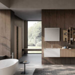 Bagno di design con basi sospese in colore beige e bianco, specchio rettangolare e pensili con aperture a giorno abbinati alle basi in colore beige. Sulla sinistra una vasca vasca freestanding bianca e in secondo piano delle colonne beige tra le pareti rivestite in legno scuro. Sullo sfondo un’ampia vetrata con vista sul giardino.